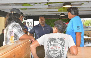 A group of young people are talking to a man in a dark blue shirt with a cross necklace. They are in a bright indoor place with green plants and wooden tables. Some people are wearing glasses and headphones. Everyone looks happy.